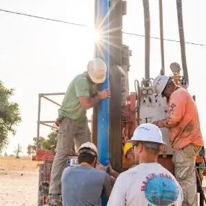Men with Friends In Action International watch as drilling rig digs hole for well in West Africa.